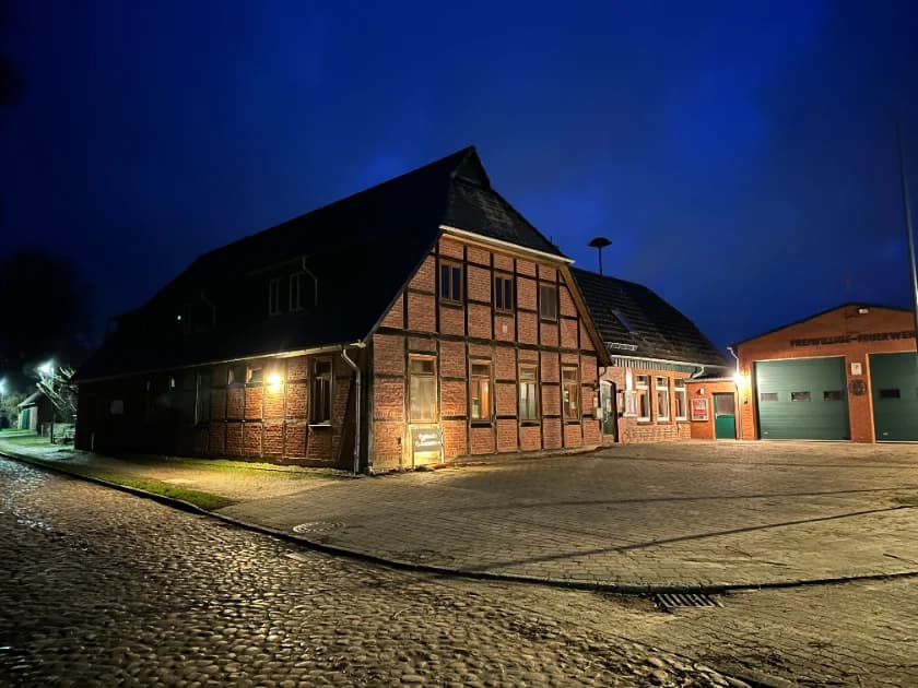 Das Kulturzentrum Witzeeze am Abend: ein rotes Fachwerkhaus mit warm erleuchteten Fenstern an der gepflasterten Dorfstraße, rechts daneben das Gerätehaus der Freiwilligen Feuerwehr